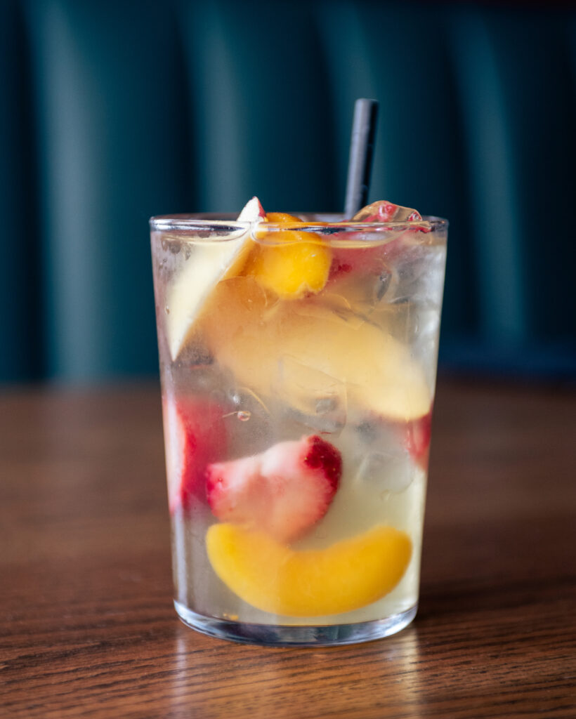 White sangria in glass with strawberries, peaches, and ice on a table with window light